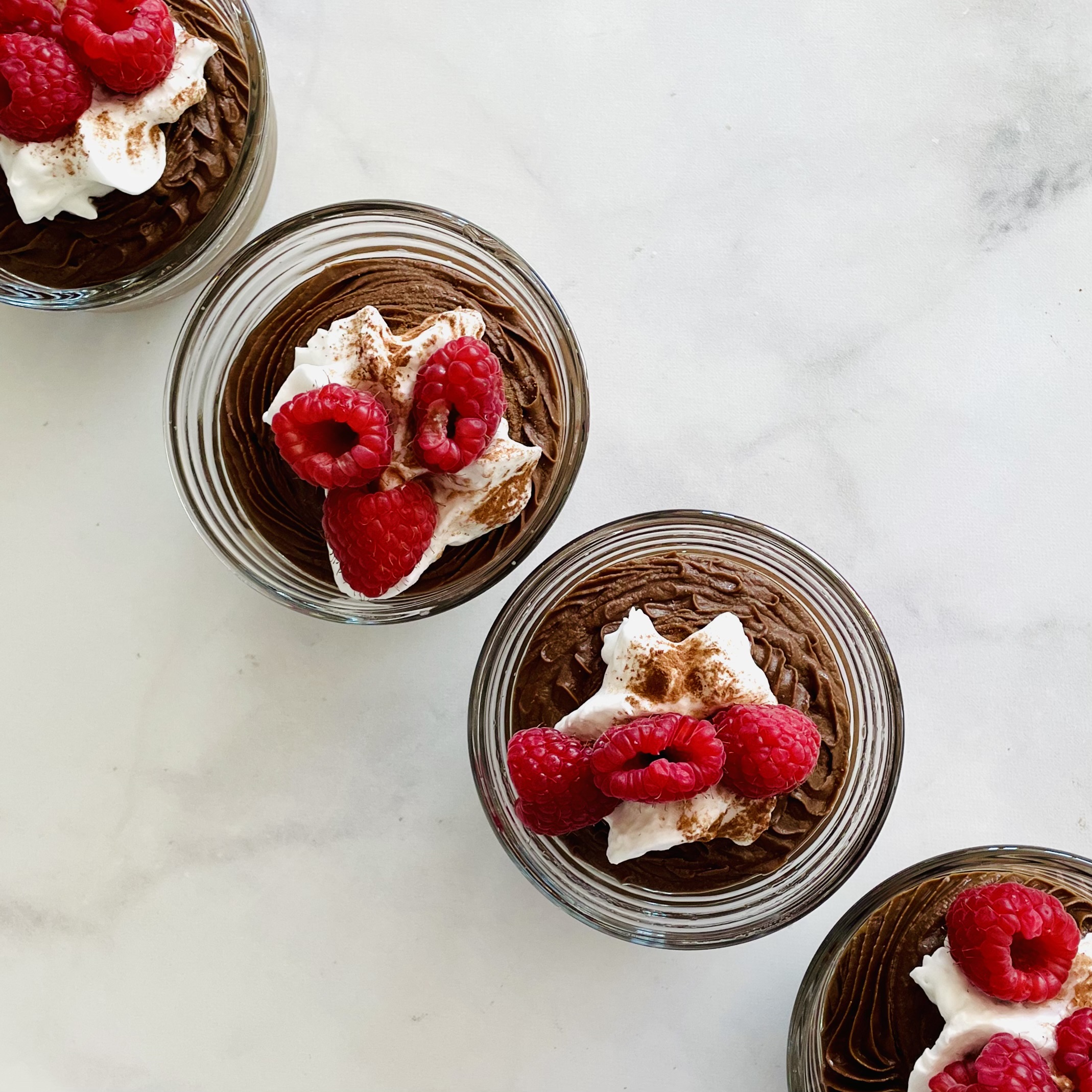 four dessert dishes with no-cook plant-based cocoa mousse toped with coconut whipped cream, cocoa, and raspberries