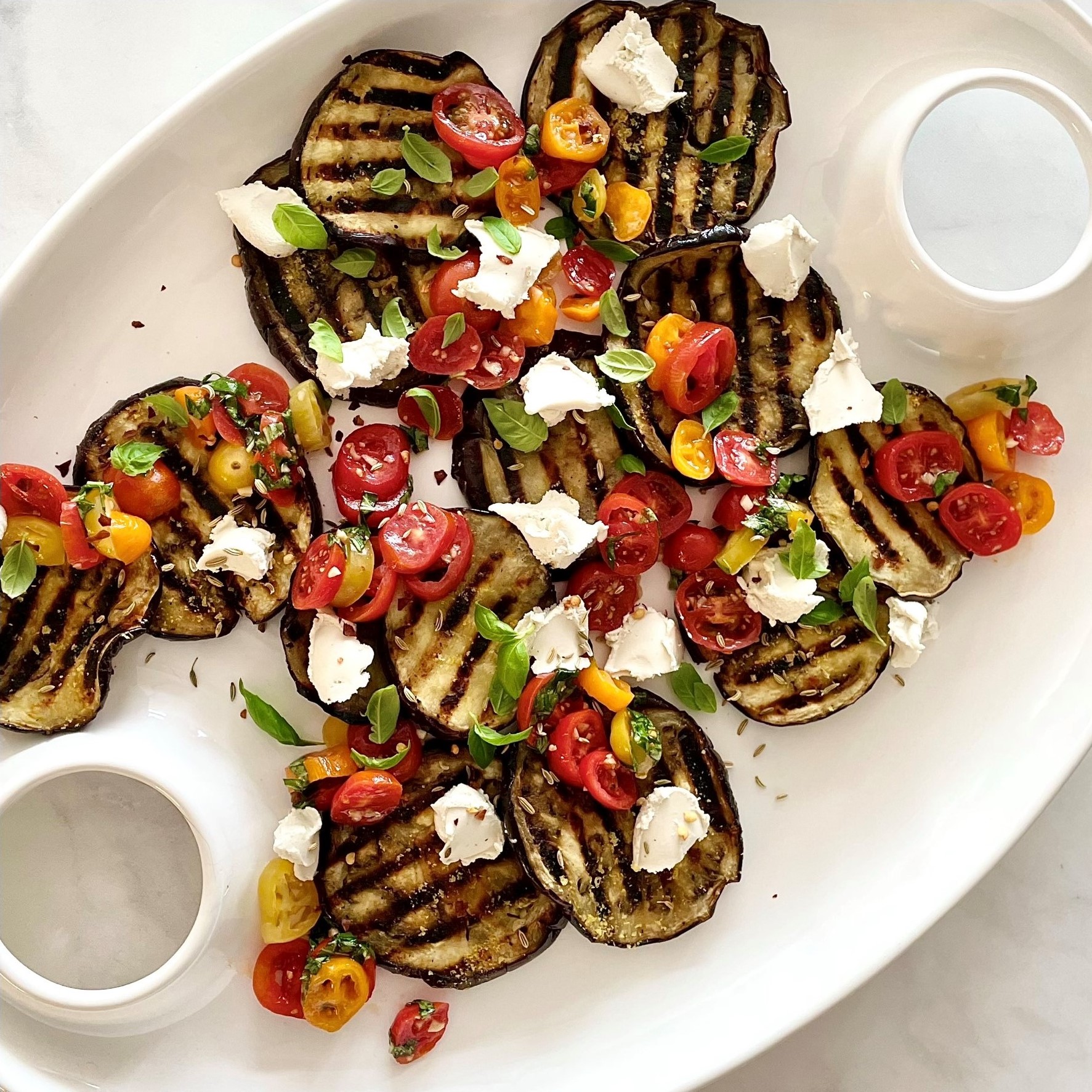 white platter of grilled eggplant carpaccio with tomato-basil salsa & treenut cheese on white marble
