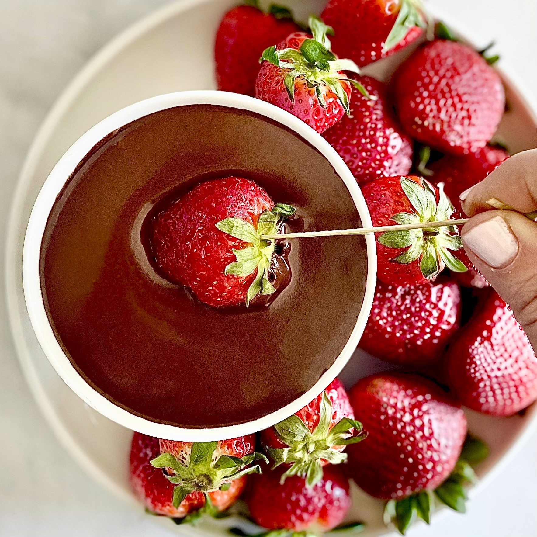 Dunking a strawberry into dark chocolate fondue sauce with plate of fresh strawberries
