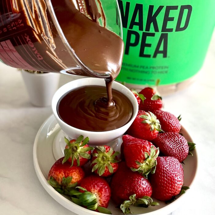 Pouring chocolate sauce into a bowl surrounded by strawberries with Naked Pea protein in background